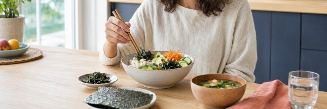 Femme dégustant un bol de riz, de légumes et d’algues comestibles dans une cuisine lumineuse.