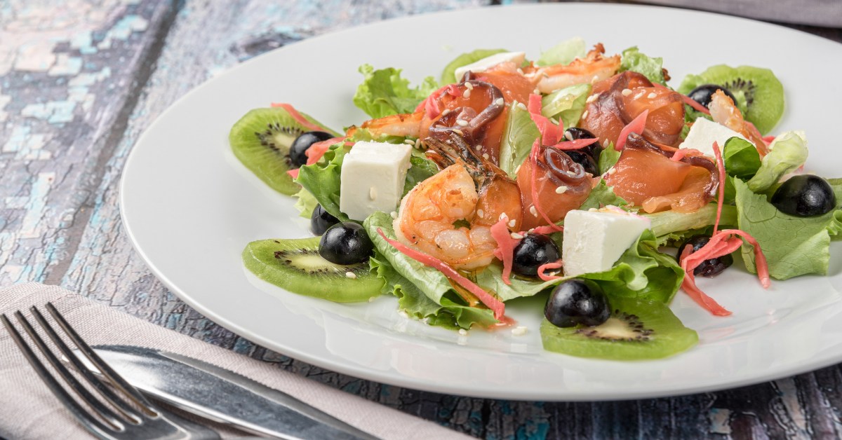 Plat de salade fraîche avec crevettes, saumon, cubes de fromage, olives, kiwi et verts mixtes.