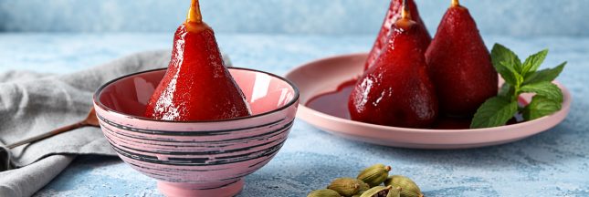 Poires pochées au vin rouge dans une vaisselle rose, avec des gousses de cardamome au premier plan.