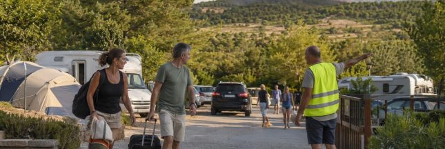 Des vacanciers quittent un camping avec leurs bagages tandis qu’un feu de forêt brûle au loin et qu’un responsable les dirige vers la sortie.