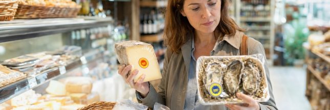 Une consommatrice compare un fromage et des huîtres portant des signes de certification dans une épicerie.