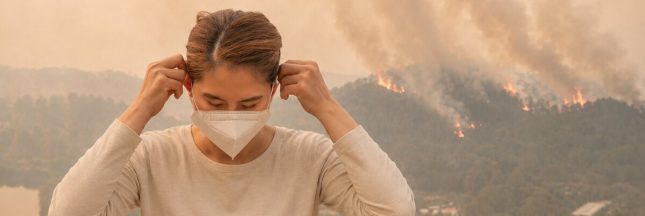 Une femme ajuste son masque tandis qu’un feu de forêt dégage d’importantes fumées sur une colline en arrière-plan.