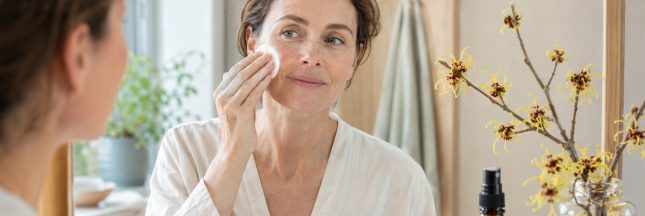 Femme appliquant un hydrolat d’hamamélis sur son visage devant un miroir, près de fleurs jaunes d’hamamélis.