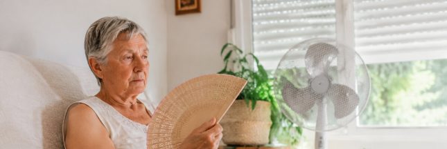 Femme âgée buvant de l’eau et utilisant un éventail devant un ventilateur pendant la canicule