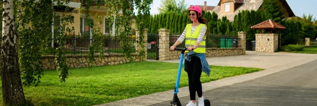 Femme portant un casque et un gilet jaune réfléchissant sur une trottinette électrique.